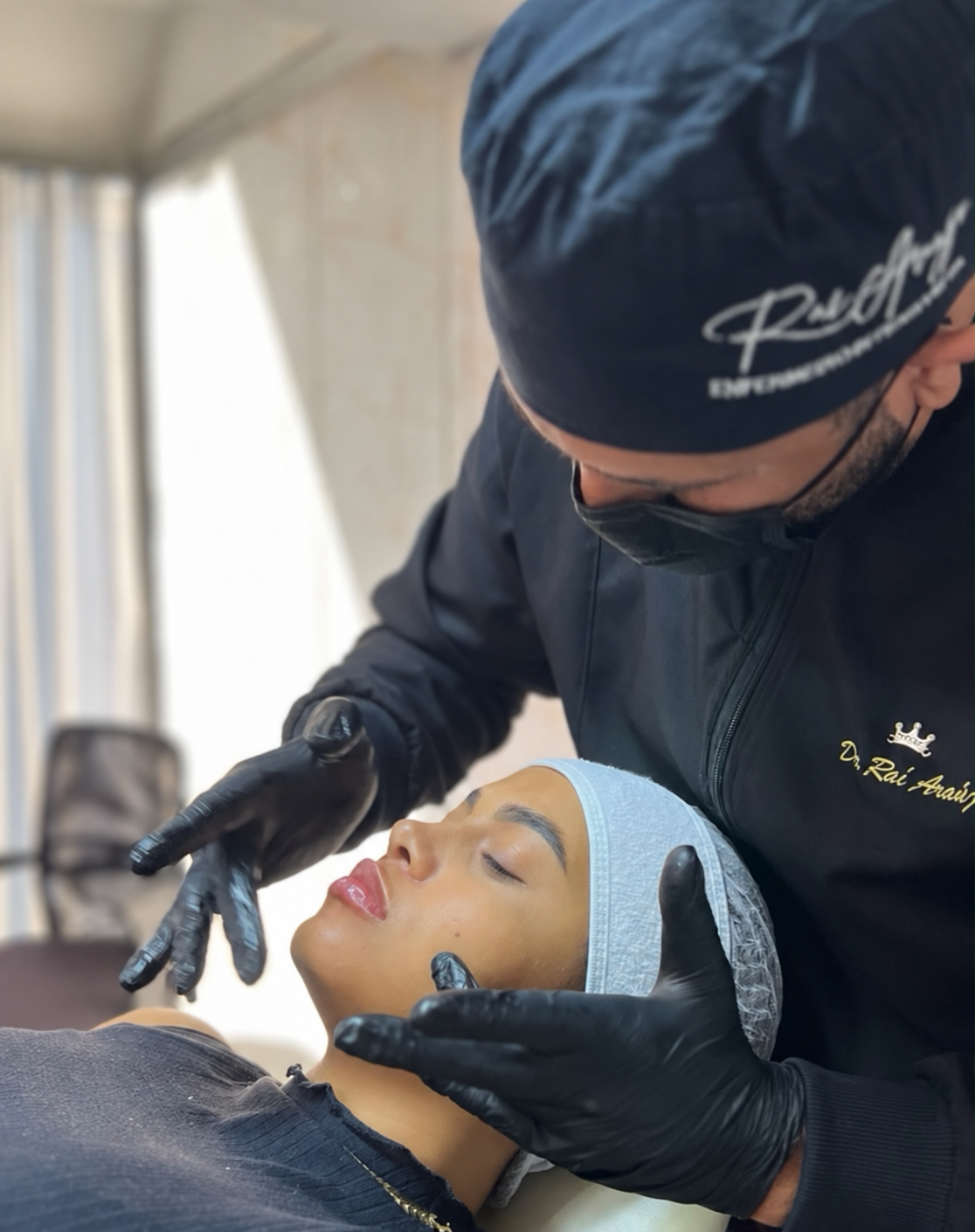 Profissional da equipe em atendimento estético facial a uma paciente deitada, com touca e máscara